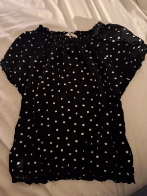 Svart prickig blus med spetsdetaljer - Söt svart blus med vita prickar och korta puffärmar. Blusen har elastisk halsringning och spetsdetaljer vid hals och ärmslut. Tillverkad i ett tunt, lätt material som ger en luftig känsla. Perfekt för dig som gillar retrovibbar och lekfull stil.