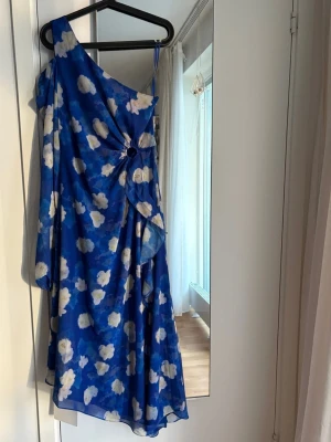 Adoore klänning strl 34 blå  - Säljer en blå one shoulder klänning med vita blommor och asymmetrisk skärning. Klänningen har en dekorativ ringdetalj framtill och är gjord i ett lätt, flygigt material som ger en snygg siluett. Perfekt för dig som vill sticka ut! Helt ny med tags! Nypris: 1695kr.