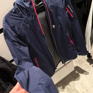 Blå vindjacka Peak Performance S - Snygg marinblå vindjacka från Peak Performance med rosa detaljer på dragkedjorna. Jackan har huva, långa ärmar och praktiska fickor med dragkedja. Perfekt för blåsiga dagar och outdoor-aktiviteter. Sportig och stilren design som funkar året runt. Säljer denna märkes jackan för 800kr men kan gå ner i pris med rimliga bud.