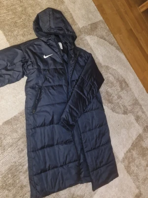 Mörkblå lång dunjacka från Nike - Snygg svart lång dunjacka från Nike med huva och dragkedja. Jackan har Therma-FIT-teknologi som håller värmen och två sidofickor med dragkedja. Perfekt för kalla vinterdagar och riktigt skön att bära. Väldigt nyligen köpt och nästan aldrig använd. Inga defekter, äkliga dofter, smutsflekar osv kommer med denna jackan. Helt ny. Storleken är S herr. Jag är personligen en väldigt biffig person och jag är 180 cm och det enda som är några centimeter för kort är ärmarna. Pris kan negotieras (snabbafär)