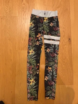 Färgglada leggings från Stronger i XS.  - Snygga leggings från stronger i XS. De har bred grå midja och två vita ränder på ena benet. Perfekta för träning och är väldigt sköna. Väl använda men hela och inga hål. 