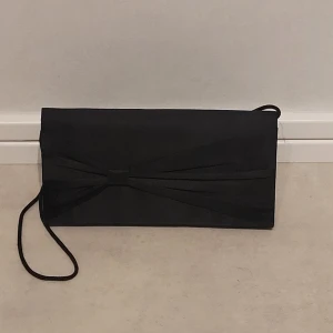 Svart kuvertväska med rosettdetalj - Stilren svart kuvertväska med elegant rosettdetalj på framsidan och tunn axelrem. Väskan har en klassisk rektangulär form och är tillverkad i ett tygmaterial med matt finish. Perfekt för dig som vill ha en enkel men snygg accessoar till din outfit.