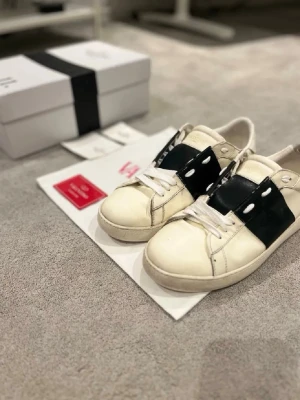 Valentino Garavani vita sneakers - Snygga vita sneakers från Valentino Garavani med bred svart läderdetalj över vristen och vita skosnören. Klassisk låg modell med rund tå och diskreta nitar på sidorna. Kommer med originalkartong och dustbag.