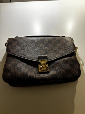 Brun axelremsväska - Snygg handväska från Louis Vuitton i klassiskt brunt damier-mönster. Väskan har guldfärgat lås och detaljer, samt en axelrem. Tillverkad i canvas med läderdetaljer. Perfekt för dig som gillar lyxiga accessoarer och tidlös design.