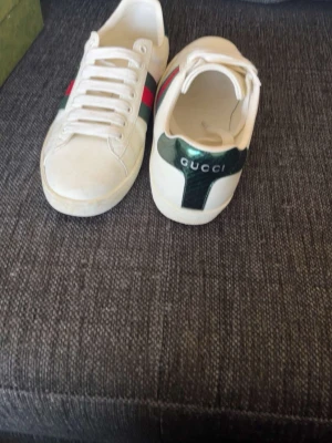 Gucci vita sneakers  - Snygga vita sneakers från Gucci med klassiska gröna och röda ränder på sidan samt metallicgrön häl med logotyp. Lågt skaft och vita snören. Tillverkade i läder för en lyxig känsla och stilren look. Kan ta mer bilder box och allt följer med. Priset ej hugget i sten