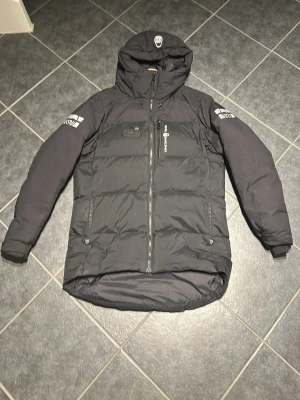 Svart pufferjacka från sail racing  - Säljer en svart pufferjacka från sail racing med huva och dragkedja framtill. Jackan har logotyp på bröstet och ärmen samt praktiska fickor med dragkedja. Perfekt för kalla vinterdagar och riktigt skön passform.