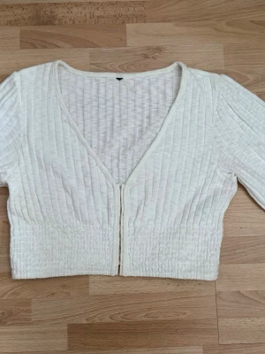 Vit croppad ribbad topp med dragkedja - Snygg vit croppad topp med ribbad struktur och v-ringning. Toppen har lång ärm och dragkedja framtill, vilket ger en modern och fräsch look. Perfekt att styla med höga jeans eller kjol. Hade på mig en gång. 