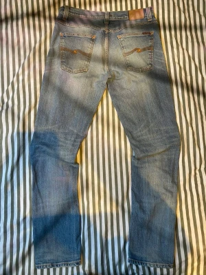 Blå jeans från Nudie Jeans Co - Säljer ett par klassiska blå jeans från Nudie Jeans Co med snygga kontrastsömmar och femficksmodell. Jeansen har en rak passform och är tillverkade i slitstark denim. Perfekta för dig som gillar tidlös och avslappnad stil.