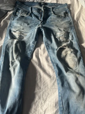 Ljusblå slitna jeans från Replay - Säljer ett par ljusblå jeans från Replay med flera slitningar och hål på framsidan. Jeansen har klassisk femficksmodell, normal midja och raka ben. Materialet är mjukt denim med snygg tvätt och slitna detaljer för en trendig look.