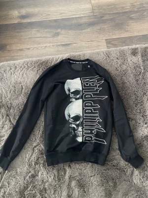  Philipp Plein  - Cool svart sweatshirt från Philipp Plein med stort tryck av två dödskallar och logotypen PHILIPP PLEIN i vitt framtill. Tröjan har rund hals, långa ärmar och ribbade muddar. Materialet känns mjukt och bekvämt, perfekt för en edgy streetwear-look.