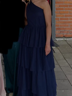 Balklänning  one shoulder volangklänning - Säljer min balklänning, en lång mörkblå klänning med one shoulder-design och flera lager volanger. Klänningen har en elegant och flytande siluett, perfekt till balen. Den är insydd men går att spreta jättelätt och kan bli storlek 36 