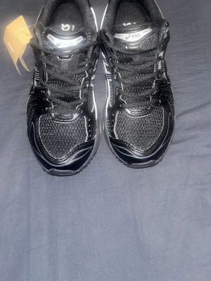 Svarta Asics sneakers med silvriga detaljer - Snygga svarta sneakers från Asics med silvriga detaljer och logga på sidan. Skorna har en sportig design med mesh och syntetmaterial för bra andningsförmåga. Klassisk snörning och bekväm sula som passar perfekt till streetwear eller träning.