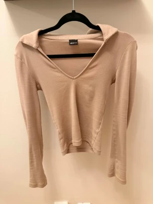 Beige långärmad topp från Gina Tricot - Trendig beige långärmad topp från Gina Tricot med v-ringning och krage. Toppens material känns mjukt och stretchigt, perfekt för en stilren look. Passformen är normal och toppen har en enkel, clean design utan mönster.