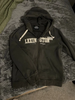 Mörkgrå Lexington hoodie med dragkedja - Snygg mörkgrå hoodie från Lexington med vit logga på bröstet och dragkedja framtill. Har huva med vita snören och två fickor. Perfekt för en chill och avslappnad stil. Mjuk insida för extra komfort.