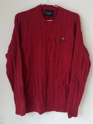 Röd Stickad Sweatshirt Bondelid - Snygg röd kabelstickad tröja från Bondelid med rund halsringning och diskret broderad logga på bröstet. Tröjan har långa ärmar och ribbade muddar vid ärmslut och nederkant. Perfekt för dig som gillar klassisk stil med en modern touch.