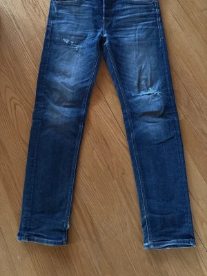Jack & Jones Blå slitna jeans med raka ben tapered/mike - Säljer ett par blå jeans med raka ben och slitningar på låren och knäna. Jeansen har klassisk femficksdesign och är gjorda i denim med snygg tvättad look. Modell tapered/mike