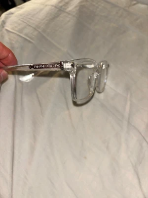 Chrome Heart glasögon  - Säljer ett par stilrena, transparenta glasögonbågar från Chrome Hearts. Bågarna har fyrkantig form och detaljer i metall på skalmarna samt en liten dekorativ blomma i hörnet. Perfekt accessoar för dig som vill sticka ut med en unik look.