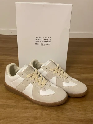 Maison Margiela gats - Säljer ett par stilrena sneakers från Maison Margiela i vitt och beige. Skorna har ovandel i slätt läder och mocka, med beigea detaljer och klassisk gummisula. Perfekt för dig som gillar minimalistisk och trendig design.  F@k3.