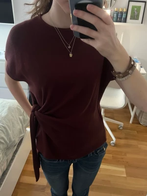 Vinröd topp med knyt i sidan - Snygg vinröd topp med korta ärmar och rund halsringning. Toppen har en cool knytdetalj i sidan som ger en unik look. Perfekt att styla med jeans eller kjol för en trendig outfit. Använt den ett fåtal gånger 🥰