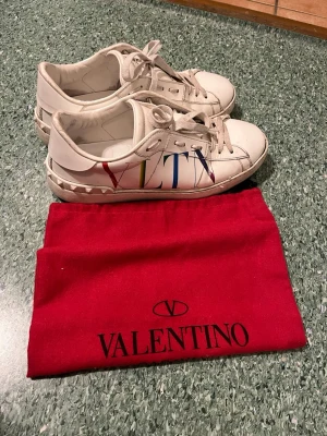 Valentino open 41 vita sneakers med VLTN-logga - Snygga vita sneakers från Valentino med färgglad VLTN-logga på sidan. Skorna har nitar på hälen och är gjorda i läder med vita snören. Kommer med röd dustbag med Valentino-tryck. Har varit mina daylies länge men nu har dem varit oanvända nåt år men de finns mycket liv kvar i dessa.
