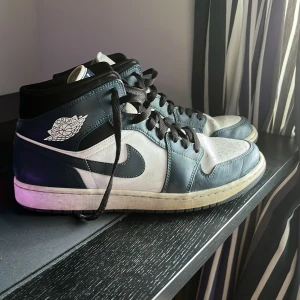 Nike Air Jordan 1 Mid Marinblå/vit - Snygga Nike Air Jordan 1 Mid sneakers i svart och vitt läder med klassisk vinge-logga på sidan och marinblå swoosh. Skorna har svarta skosnören och vit mellansula. Perfekta för dig som gillar streetwear och vill ha en ikonisk look.