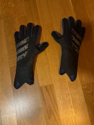 Svarta Adidas målvaktshandskar - Svarta målvaktshandskar från Adidas med gråa ränder på ovansidan och blå detaljer. Handskarna har en greppig yta och lång mudd för extra stöd. Perfekta för dig som spelar fotboll och vill ha bra grepp och stil på planen. Hör av er privat om ni har några frågor