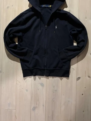 Svart hoodie från Polo Ralph Lauren - Hoddie från Polo Ralph Lauren. Klassisk design med huva och dragsnören, två fickor framtill och den ikoniska broderade loggan på bröstet. Tillverkad i mjukt bomullsmaterial som är skönt att bära.
