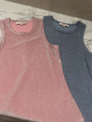 Glittrig rosa linnetopp från A View - Supersnygg rosa linnetopp från A View med glittrigt material som verkligen fångar ljuset. Ärmlös modell med rund halsringning och en mjuk, stretchig känsla. Perfekt för dig som vill sticka ut lite extra.
