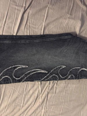 Svarta jeans med våg- och flamebrodyr - Unika svarta jeans med grå tvätt och broderade vågor och flames längs benen. Raka ben och klassisk femficksmodell. Perfekta för dig som vill sticka ut med coola detaljer. Materialet är denim, troligtvis bomull.