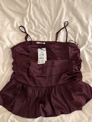Mörkröd satinlinnetopp Gina Tricot - Supersnygg mörkröd satinlinnetopp från Gina Tricot med tunna axelband och rynkade sidor. Toppen har en volang nedtill som ger en extra touch och ett glansigt material som känns lyxigt. Perfekt att styla med jeans eller kjol.