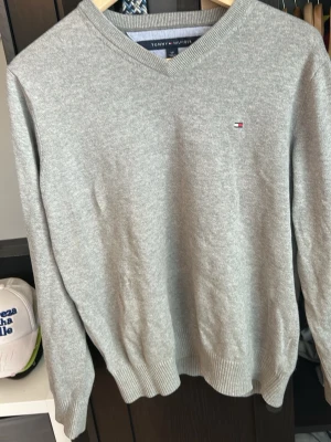 Grå stickad tröja Tommy Hilfiger - Snygg grå stickad tröja från Tommy Hilfiger med V-ringning och diskret logga broderad på bröstet. Tröjan har ribbade muddar vid ärmslut och nederkant. Perfekt till jeans eller chinos för en clean look.