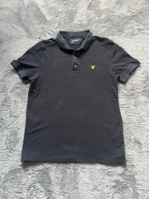 Svart pikétröja från Lyle & Scott - Klassisk svart pikétröja från Lyle & Scott med korta ärmar och gul logga på bröstet. Tröjan har krage och två knappar framtill. Tillverkad i mjuk bomull som känns skön mot huden. Perfekt till jeans eller shorts för en clean look.