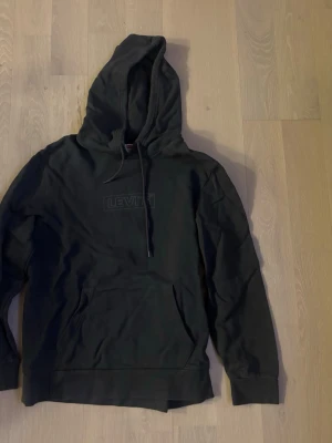 Svart hoodie från Levi's med logga - Snygg svart hoodie från Levi's med diskret broderad logga på bröstet. Klassisk känguruficka framtill och justerbar huva med dragsko. Tillverkad i mjuk bomullsmix som är skön att ha på sig. Perfekt för en avslappnad och cool stil.