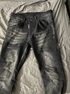 Svarta Dsquared2 jeans med färgstänk - Säljer ett par svarta jeans från Dsquared2 med coola slitningar och färgstänk i blått och vitt. Jeansen har fem fickor, knappgylf och en snygg tvättad look. Perfekta för dig som gillar streetstyle och unika detaljer.