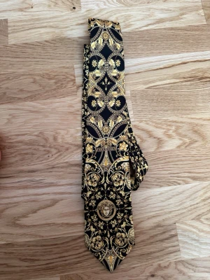 Versace slips – svart/guld - Stilren slips i svart med detaljerat guldmönster och barockinspirerade motiv. Slipsen har klassisk form och är dekorerad med blommor, ornament och ett ansikte i mitten. Materialet ser ut att vara polyester eller siden för en lyxig känsla.