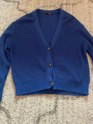 Blå stickad kofta med knappar - Snygg blå stickad kofta med tre knappar framtill och v-ringad hals. Kortare modell med ribbade muddar vid ärmslut och nederkant. Perfekt att slänga över en t-shirt eller linne för en chill look.