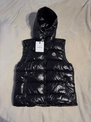 Svart dunväst från Moncler med huva - Snygg svart dunväst från Moncler med glansig finish och huva. Västen har dragkedja framtill, två fickor med dragkedja och Moncler-logga på bröstet. Perfekt för lager-på-lager och streetstyle.