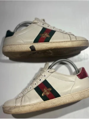 Gucci vita sneakers med bi-detalj - Snygga vita sneakers från Gucci med klassiska gröna och röda ränder på sidan samt en guldfärgad bi-detalj. Skorna har lågt skaft, vit sula och röda samt gröna detaljer på hälen. Tillverkade i läder med snörning framtill. Den svarta klumpen e en lera som jag redan har tagit bort 