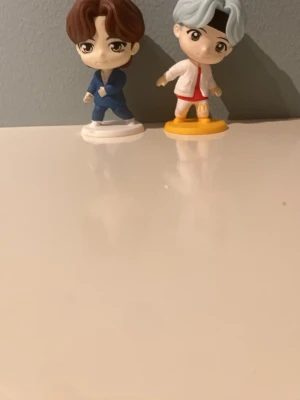 Två anime-inspirerade samlarfigurer - Säljer två små samlarfigurer i plast med anime-stil. Ena figuren har brunt hår och blå kostym, den andra har ljusblått hår, vit jacka och röda shorts samt står på en gul bas. Perfekt för dig som gillar manga eller vill dekorera skrivbordet.