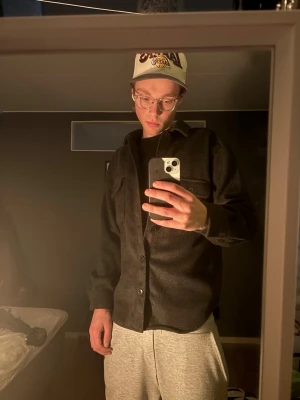 Mörkgrå overshirt med ficka - Säljer en stilren mörkgrå overshirt med knäppning framtill och bröstficka med lock. Overshirten har en rak passform och är perfekt att bära över en t-shirt. Materialet ser ut att vara ullblandning eller liknande för en mjuk känsla.