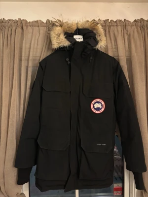 Svart Jacka från Canada Goose - Säljer en svart jacka från Canada Goose med klassisk patch på bröstet. Jackan har stor huva med fuskpäls, fyra stora fickor framtill och dragkedja med vindslå. Perfekt för kalla dagar och riktigt snygg streetstil.