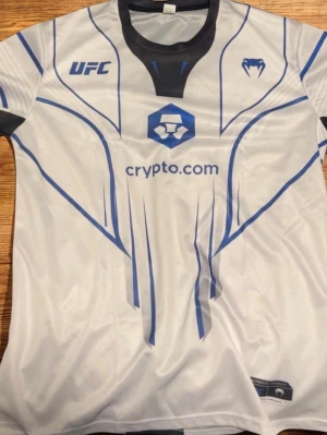 UFC x Venum vit sporttröja - Snygg vit sporttröja från Venum i samarbete med UFC. Tröjan har blå och svarta grafiska linjer, svart krage och detaljer samt tryck från crypto.com. Perfekt för träning eller att bära som supporter. Materialet är lätt och andas. KAN KÄNNAS MINDRE FÖR DEN ÄR COMPRESSED