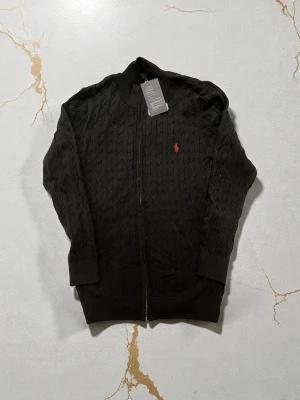 Svart kabelstickad ziptröja Polo Ralph Lauren - Svart kabelstickad ziptröja från Polo Ralph Lauren med röd broderad logga på bröstet. Tröjan har ribbade muddar, hög krage och dragkedja framtill. Perfekt för en clean och stilren look.