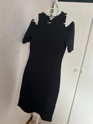 Svart bodycon klänning med cut-outs - Snygg svart bodycon klänning med trendiga cut-outs vid axlarna och korta ärmar. Klänningen har en enkel, tight passform och är gjord i ett stretchigt material som sitter bekvämt. Perfekt för dig som gillar stilrena och moderna plagg.
