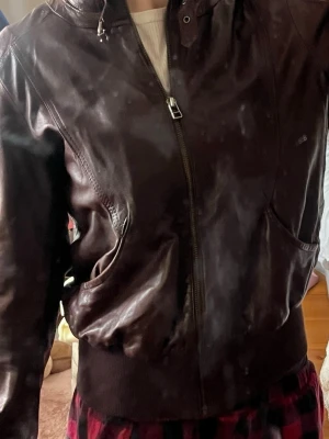 Mörkbrun skinnjacka med dragkedja - Snygg mörkbrun skinnjacka med dragkedja framtill och två snedställda fickor. Den är väldigt liten i storleken. Jackan har ribbade muddar vid midja och ärmslut för en tajtare passform. Klassisk modell som ger en cool look och passar perfekt till många olika stilar.