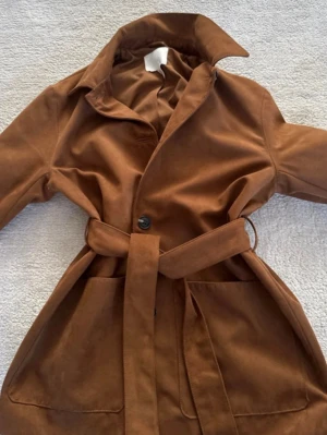 Brun trenchcoat med knytskärp - Snygg brun trenchcoat från H&M med klassisk krage, stora framfickor och knytskärp i midjan. Jackan har en stilren design med en knapp framtill och är tillverkad i ett mjukt tyg som ger en avslappnad men trendig look.