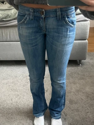 Blå bootcut jeans med fickdetaljer - Snygga blå bootcut jeans från miss sixty med ljusa slitningar och coola fickdetaljer bak. Jeansen har klassisk femficksdesign, låg midja och stängs med knapp och dragkedja. Perfekta för en avslappnad och trendig look. Size 26