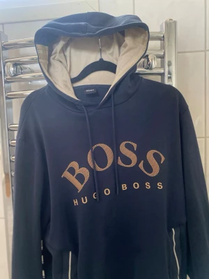 Marinblå hoodie från Hugo Boss - Snygg marinblå hoodie från Hugo Boss med stor logga i beige framtill. Hoodien har dragsko i huvan, kontrastfärgad insida på huvan och praktisk magficka. Tillverkad i mjukt material som känns skönt mot huden. Perfekt för en avslappnad och trendig look. 