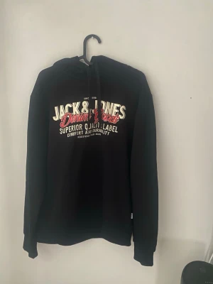 Svart hoodie från Jack & Jones - Snygg svart hoodie från Jack & Jones med tryck i vitt och rött på bröstet där det står 'Jack & Jones Denim Goods'. Hoodien har huva. Skriv för mått. Pris kan diskuteras.