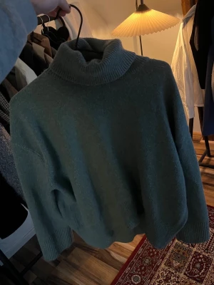 Grön polotröja i stickat material - Mysig stickad polotröja i en dämpad grön färg. Tröjan har en hög, ribbad polokrage och långa ärmar med ribbade muddar. Passformen är något oversized och materialet är mjukt och varmt, perfekt för kyliga dagar.